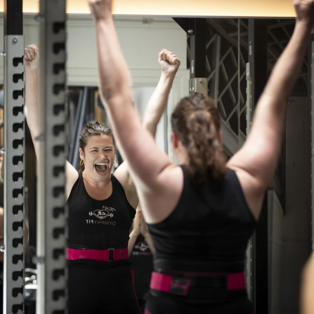 Client Tess raises arms and cheers after setting a personal record of a deadlift of 122 kilo.